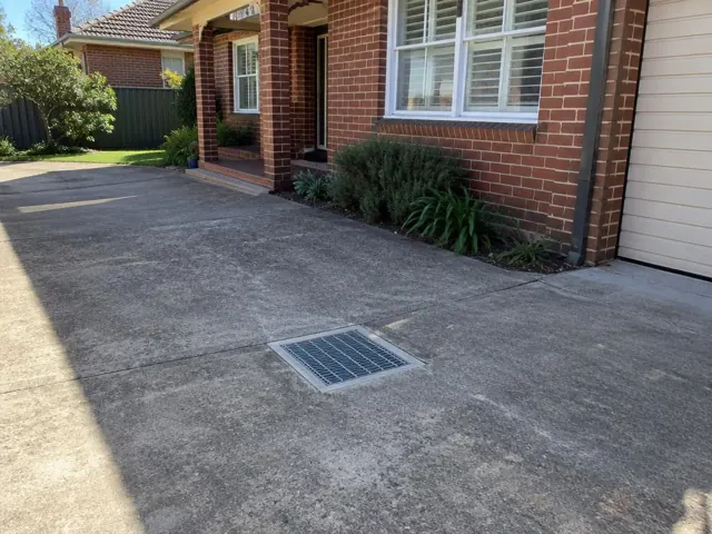 Clean outdoor drain grate set in a concrete driveway beside a brick house in Croydon